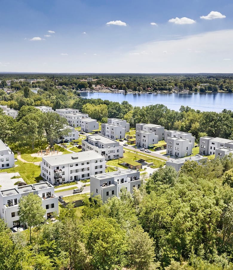 Luftaufnahme weißer kubischer Stadthäuser mit Gärten nahe einem See und Wald