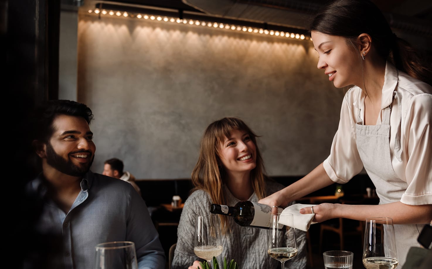 Lächelnde Kellnerin schenkt zwei fröhlichen Gästen im Restaurant Weißwein ein