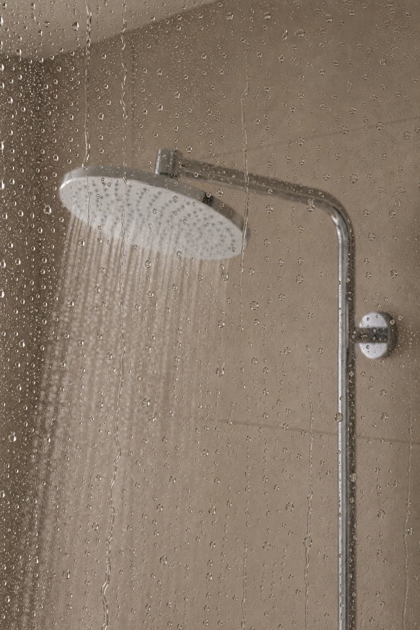 Regendusche mit Wasserstrahl vor beiger Fliesenwand hinter beschlagener Glasscheibe