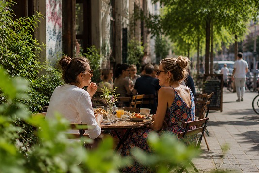 Zwei Frauen frühstücken an einem Tisch auf dem sonnigen Bürgersteig zwischen grünen Pflanzen
