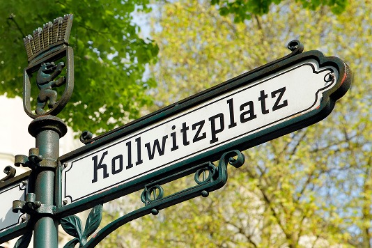 Gusseisernes Straßenschild Kollwitzplatz mit Berliner Bärenwappen vor frühlingsgrünen Bäumen