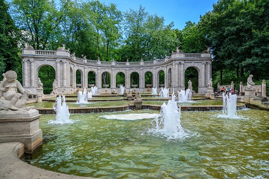 Märchenbrunnen mit Wasserfontänen, Skulpturen und steinerner Arkade in einem grünen Park