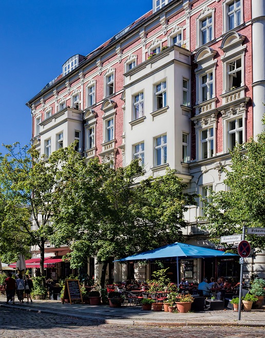 Rot-weiße Altbau-Eckfassade mit Straßencafé unter blauem Sonnenschirm und Bäumen