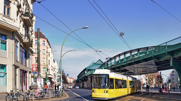 Gelbe Straßenbahn fährt bei Sonnenschein unter einer grünen Hochbahnbrücke hindurch