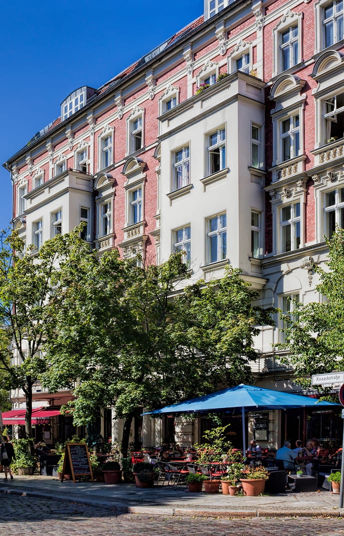 Gründerzeit-Altbaufassade mit Straßencafé unter Sonnenschirm zwischen grünen Bäumen