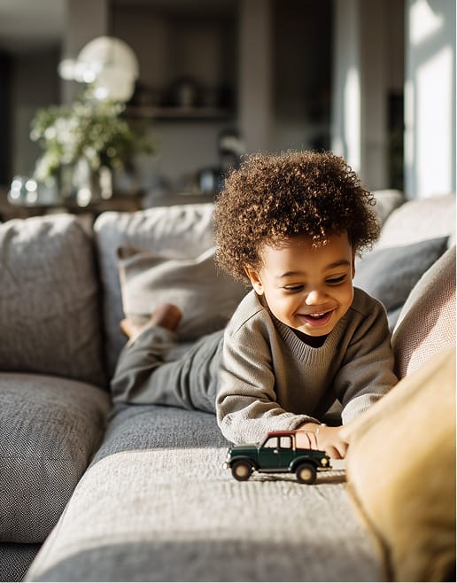 Lachendes Kleinkind liegt auf einem grauen Sofa und spielt mit einem gruenen Spielzeugauto